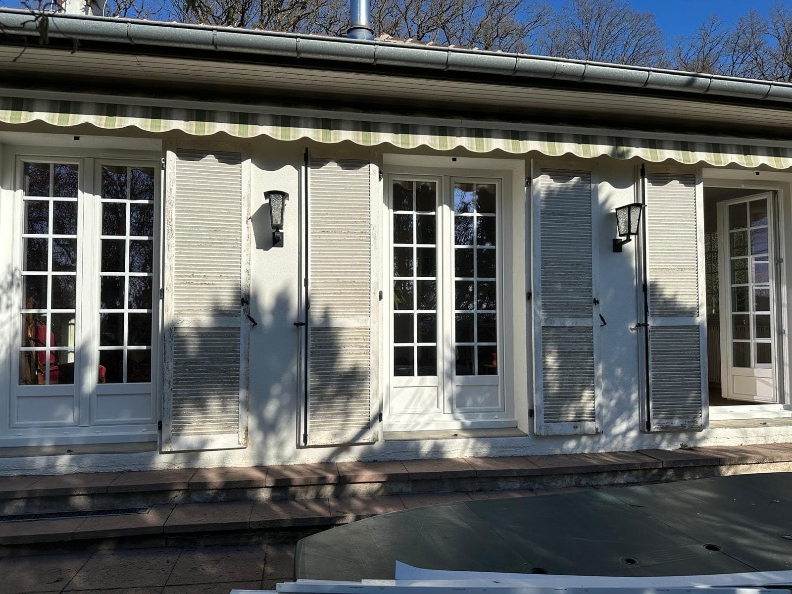 Terrasse couverte avec portes-fenêtres PVC à petits-bois et volets battants sous une avancée de toit, par REM Fermetures
